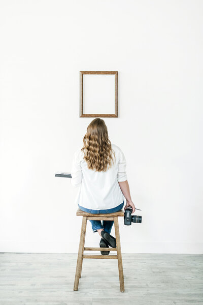 woman sitting on a wooden stool looking at a blank frame on a white wall