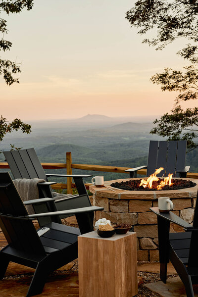 A scenic outdoor patio with black chairs surrounding a stone fire pit. Two mugs and a bowl are on a wooden table. Trees frame a view of rolling hills and a distant mountain under a softly lit sky.