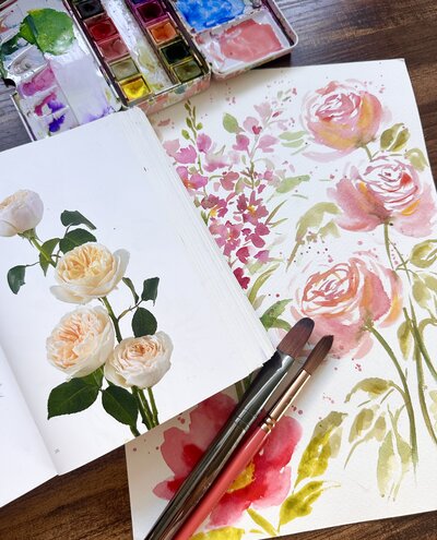 Watercolor rose painting on a table with paint brushes and a flower book and paint palette