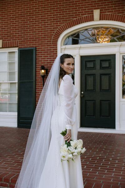 The bride is walking up the stairs of a manor holing her bouquet looking back at the camera.