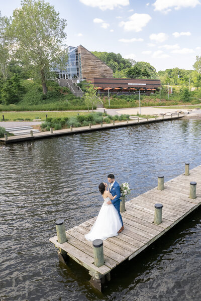 A bride and groom embrace on a wooden dock by the water, with a modern building and lush greenery in the background. The bride holds a bouquet, and the sky is partly cloudy.