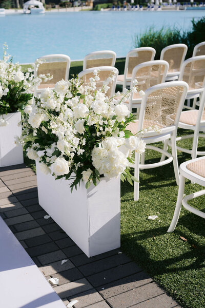 White cane-back chairs with a white floral planter beside blue water at an outdoor Houston ceremony.