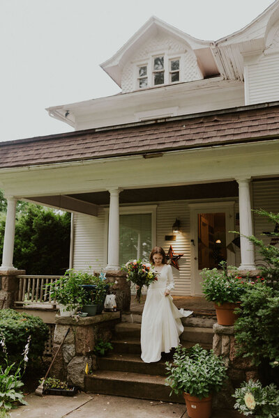 Bride walking down front porch steps in front of a tan farmhouse style home holding her dress and bouquet
