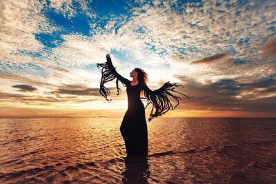 Photo of an Eclectic witch in a long black dress with long fringed sleeves dancing at the water's edge beneath a beautiful blue sky with clouds