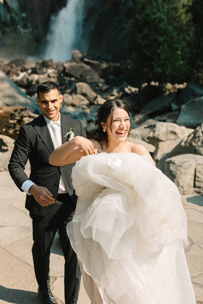 Bride laughing while groom follows behind her during their Yosemite Elopement at Lower Yosemite falls