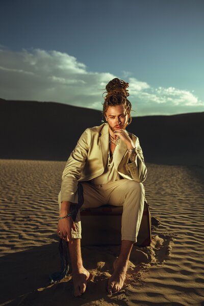 blue sky with clouds, sand hills, brown luggage, cream suit, man bun, facial hair, man sitting on luggage