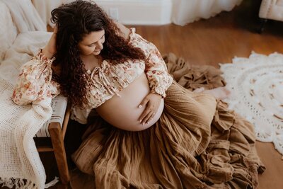 Pregnant woman with curly hair, in a floral top and brown skirt, sits on a couch, smiling and touching her belly, surrounded by cozy decor and soft lighting.