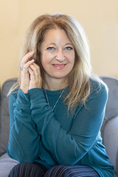 Cyndi Stewart smiling woman with long, wavy hair sits comfortably on a couch, wearing a teal top and jewelry, in a welcoming and relaxed setting.