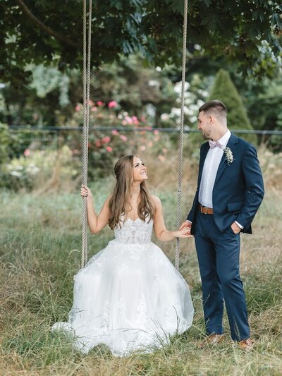 Bride sitting on a swing and smiling at groom.
