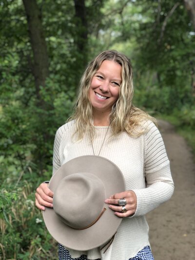 Andrea wearing a creme shirt and cream hat in her hands smiling in nature