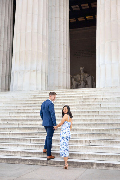 A couple holding hands and smiling at the camera while walking up the steps of the Lincoln Memorial. The man is wearing a blue suit, and the woman is in a white and blue floral dress.