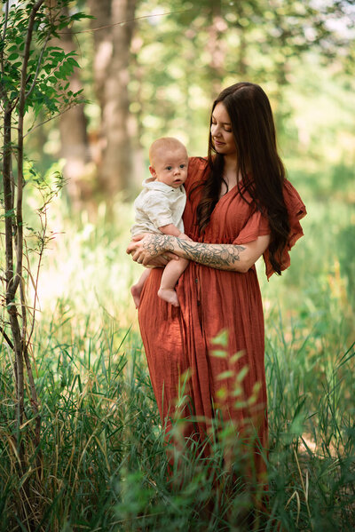 A beautiful springtime portrait of a mother with long brown hair and wearing a rust coloured dress holds her baby boy on her hip who is wearing a linen matching set. They are walking through tall green grass. Captured by Brittany Danielle in Kamloops.