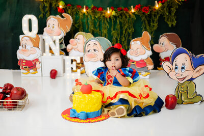 Baby in a colorful dress with a finger in her mouth, sitting next to a yellow and blue cake topped with a red apple.