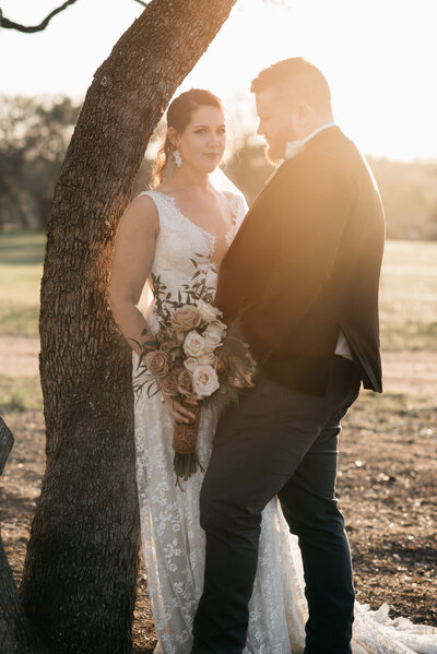 Bride leaning on tree during a portrait session with groom  admiring and sunset behind