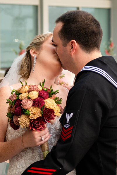 Bride and Groom kissing on a glass walkway in urban Norfolk Waterside district.  Beautiful lines and framing.  Small amount of flash light on front of Bride and Groom.  Bride holding bouquet.  Dressed in white  strappy dress with mid length veil, holding a bouquet of florals.  Groom in navy dress uniform.