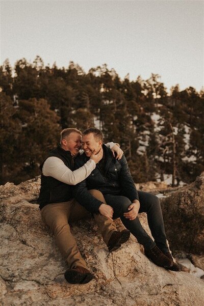 A couple shares a joyful moment sitting together on a rocky outcrop in Mount Lemmon, Tucson, Arizona. They are laughing and leaning into each other, surrounded by a backdrop of trees and evening light.