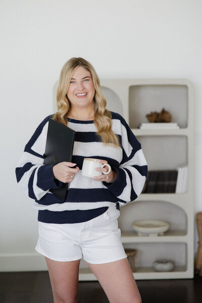 Brogan standing with a mug and laptop, wearing a cozy sweater