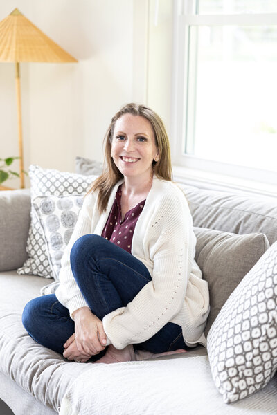 A woman with light brown hair and a warm smile sits on a gray couch, wearing a cozy white cardigan and blue jeans. She is relaxed, surrounded by patterned pillows, with natural light streaming in.