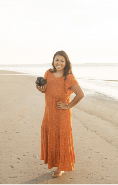 A photographer in an orange dress stands on a beach holding a camera.