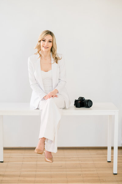 photographer sitting on white table wearing a white suit and nude heels with camera next to her