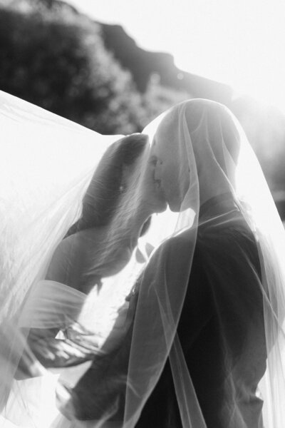 Close up in B&W of bride and groom under the veil, kissing with sunlight streaming through