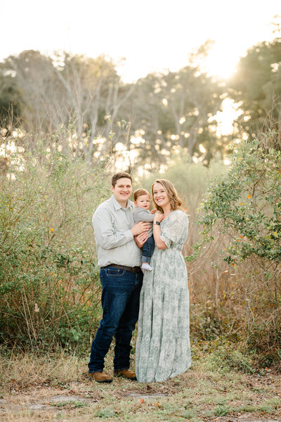 The picture captures a heartwarming family moment set against a stunning natural backdrop. The family of three - mom, dad, and their young son - stand together in a field ablaze with the vibrant hues of autumn. The lush forest in the distance provides a serene backdrop, while the warm glow of the setting sun bathes the scene in a soft, golden light.  The parents stand side by side, their son nestled comfortably between them. All three faces are alight with genuine smiles, their expressions radiating joy and contentment. The family's casual, relaxed poses suggest a sense of ease and comfort with one another, creating a sense of intimacy and connection.  The overall style of the photograph is light and airy, with a focus on capturing the natural beauty of the surroundings. The photographer, Bri Sullivan, has done an excellent job of framing the shot, ensuring that the family is the central focus while still allowing the stunning fall foliage and serene forest to shine.  The result is a beautiful, timeless portrait that perfectly encapsulates the warmth and love of a family enjoying a moment of connection in the great outdoors. It's a photograph that invites the viewer to pause and appreciate the simple pleasures of life, and the importance of cherishing those special moments with loved ones.