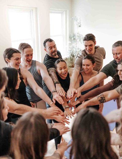 A diverse group of people in a breathwork class standing in a circle, placing their hands together in a show of unity and shared energy.