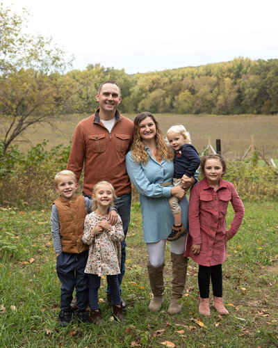 Family of 6 standing in front of a field. Mom is holding toddler boy with other children standing.