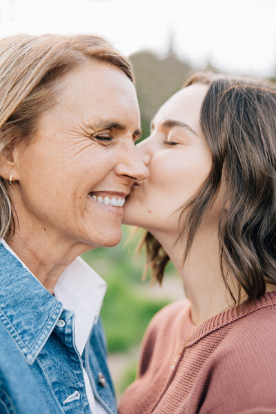 Mother and grown daughter smiling as daughter gives her a kiss on the cheek.