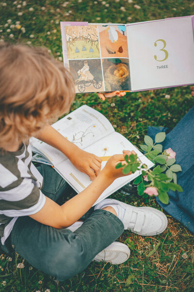 A child sitting on the grass holding a plant, engaging with an open book about nature, part of the Journeys program for ages 6-9.