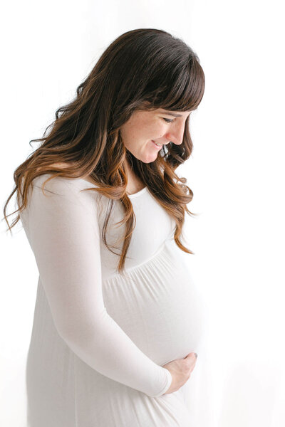 Profile image of pregnant woman in white dress holding her baby bump. She is smiling and looking down at her bump. Her hair is brown and long in loose curls falling down around her shoulders. She is backlit, so the background is white and bright.