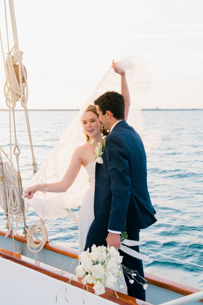 Newport Wedding Photographer | Rhode Island Caprio Captured Photography | Bride Holding Her Veil While The Groom is Holding Her Bouquet While Embracing At a Sailboat