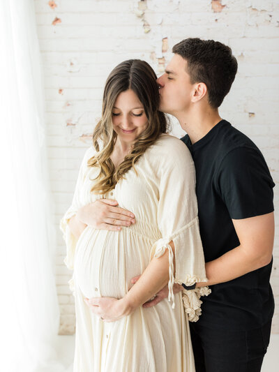 husband kissing pregnant wife in cream maternity gown for studio photoshoot cleveland maternity photographer