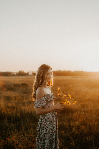 Photo of a senior girl in a field of wildflowers looking down at her freshly picked sunflowers and smiling with the sun in the background.