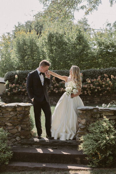 Groom kisses the bride’s hand in a sunlit garden.