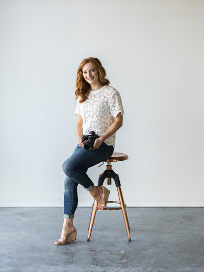 Portrait of Kansas City family photographer Katie sitting on a copper-legged stool holding her camera in a modern studio with soft lighting.
