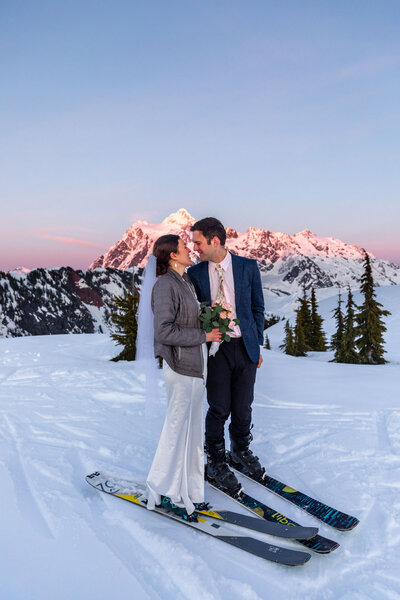 The bride and groom are wearing skis looking at each other with the mountain in the background at sunset