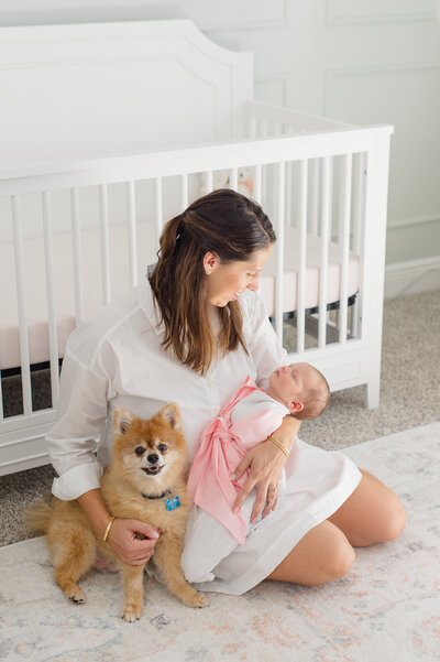 New mother sits in front of her daughters crib holding her newborn baby and dog while admiring her daughters beauty capture by a newborn photographer