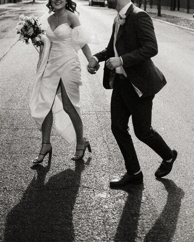 A couple joyfully runs down a street during their wedding in Chicago, Illinois. One partner is dressed in an elegant white gown with a thigh-high slit, holding a bouquet, while the other is in a suit, both captured mid-laughter in black-and-white.