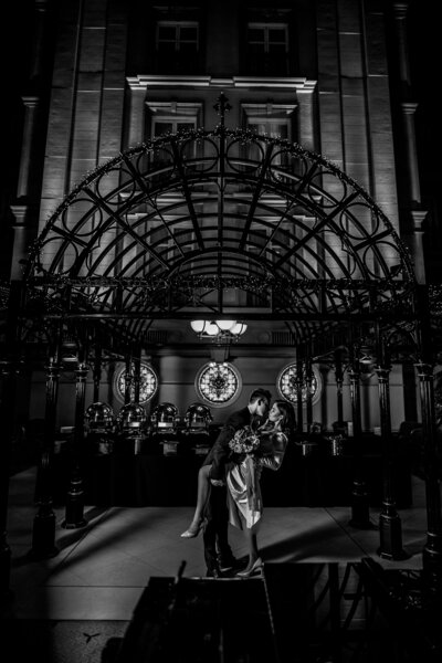 Luxurious and emotive black-and-white photo of a couple sharing an intimate moment under elegant architecture at night.