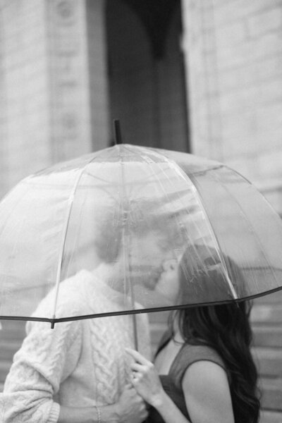A couple kisses under a clear umbrella in a filmy engagement session.