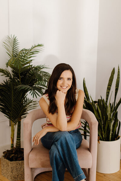 Woman with dark hair sitting in pink chair with plants behind her