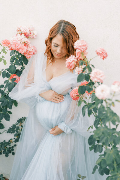 pregnant women in blue tulle dress leaning on a white wall holding her belly surrounded by pink roses