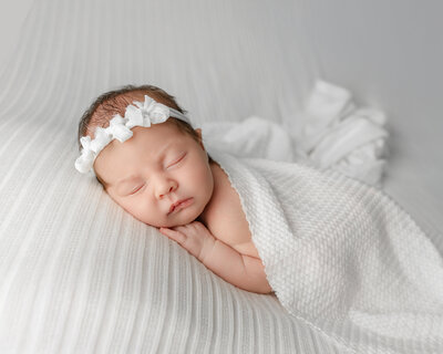 Peaceful baby girl posed in a natural side-laying position with a white bow headband and simple white tones. Photographed by Inland Empire newborn photographer Kaitlyn Dawn.