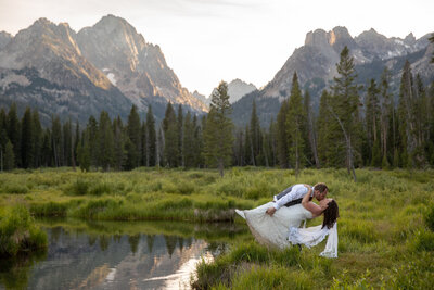 Groom dips the bride in a romantic kiss beside a reflective alpine lake, surrounded by towering jagged peaks and lush greenery.