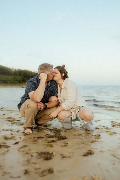 Two people kneel on a sandy beach near the water, with one person kissing the other's cheek. Both appear relaxed and happy, surrounded by shoreline and greenery in the background.