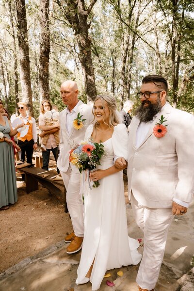 a bride walks with arms linked with dad and stepdad as they walk her down the aisle under a canopy of pine trees in greenville, texas. Captured by Shelly Voss