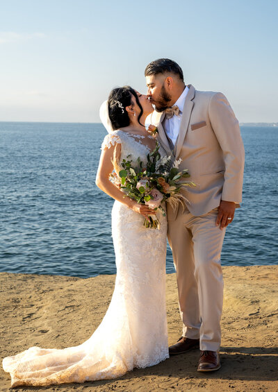 groom wearing cream suit and bride wearing white gown holding flowers kiss at their beach cliff wedding ceremony