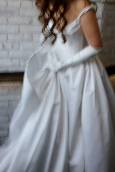 April M Payne Photography captured a soft, blurred shot of a bride in a stunning white gown with satin fabric and long gloves, her back turned to the camera, with a textured white wall in the background.