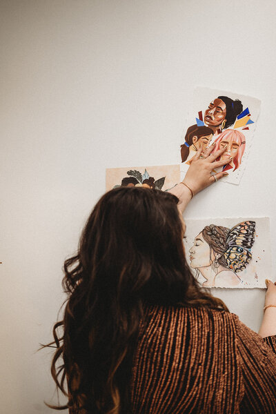 Hair stylist with long brunette waves curating a beauty inspiration wall at Haven Beauty Collective salon.